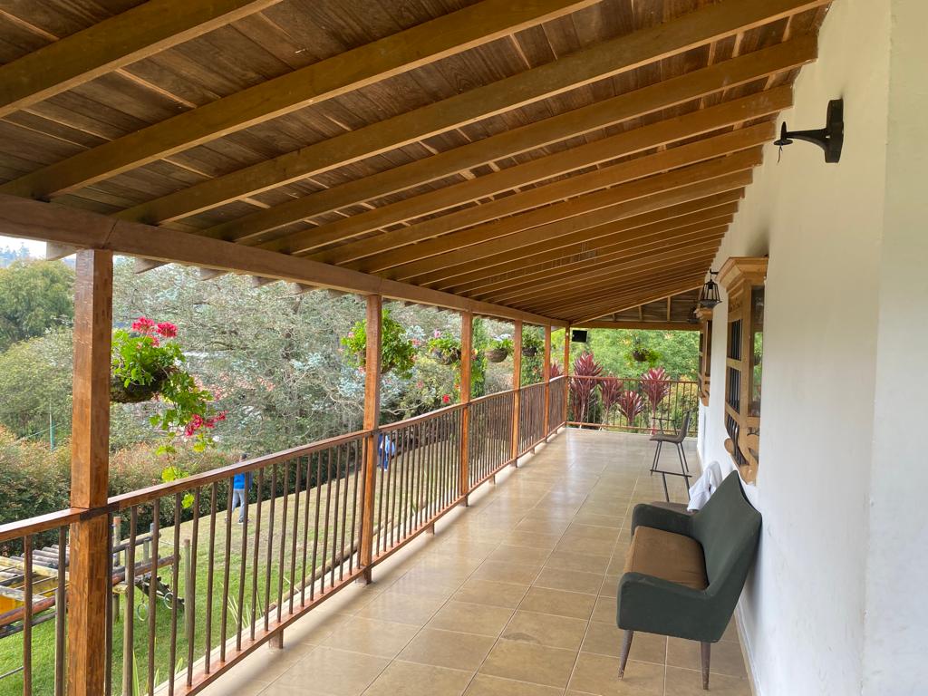 Amplio corredor con techo de madera, barandas, macetas colgantes y una banca, con vista a los jardines