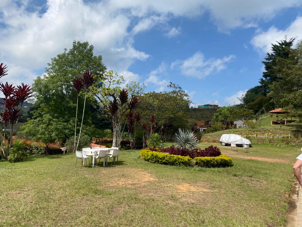 Zona verde con una mesa y sillas blancas, un macizo de flores y un kiosco al fondo