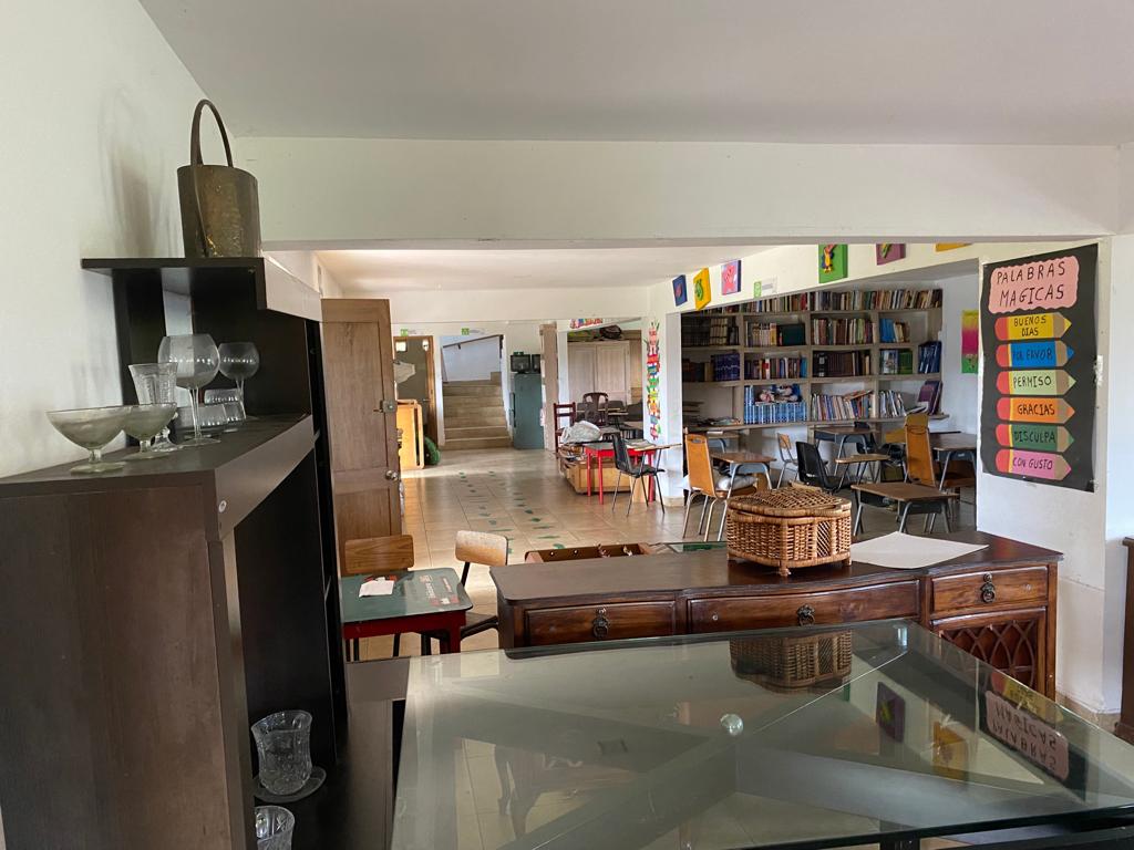 Interior de una sala con biblioteca, mesas, pupitres, un mueble de madera y un cartel de «Palabras mágicas»