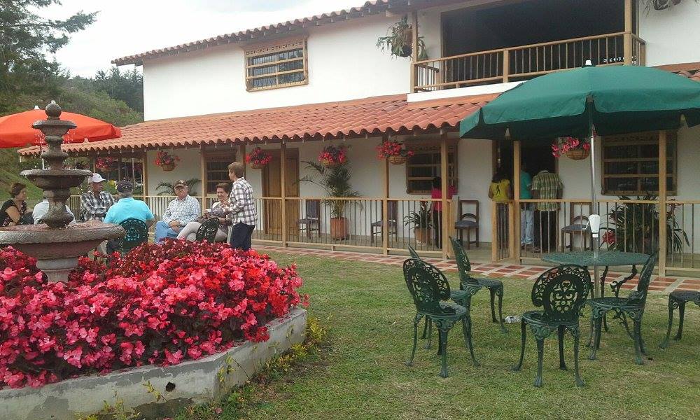 Casa principal de dos pisos con corredor, jardinera de flores rojas, fuente y mesas con sillas donde varias personas conversan