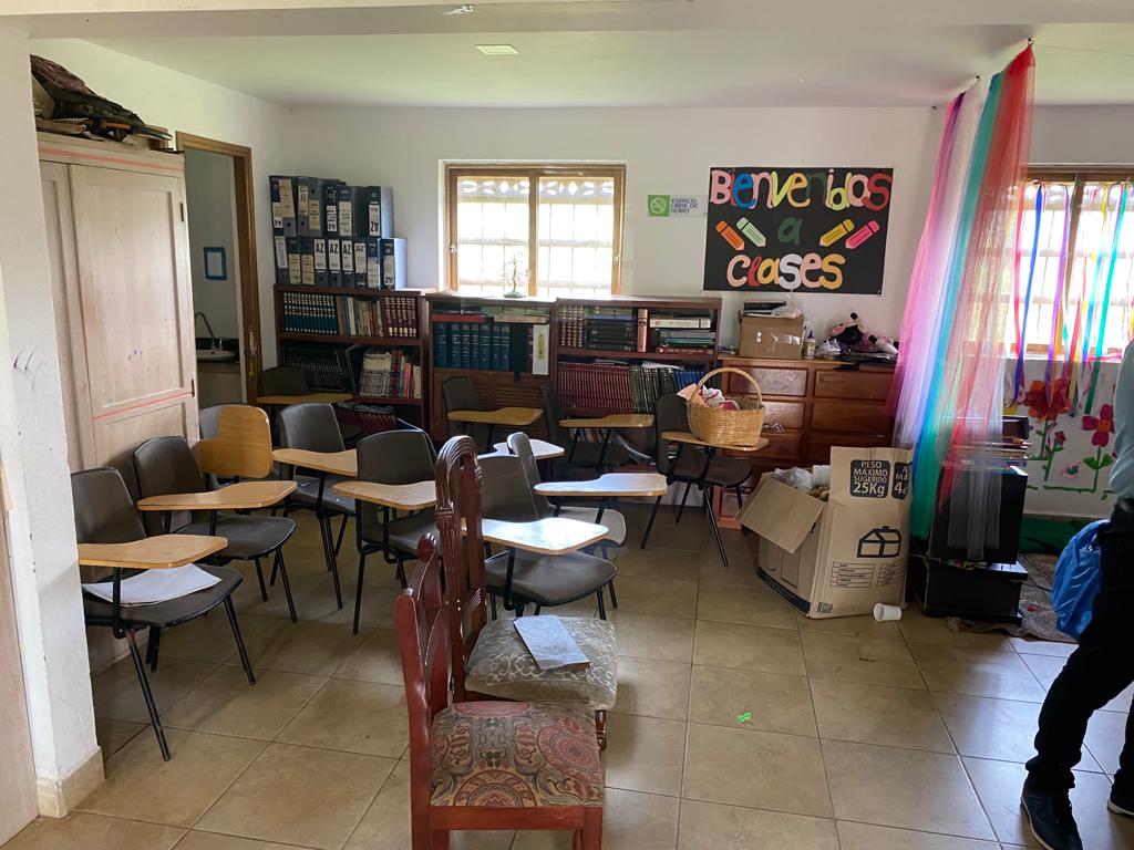 Salón con pupitres, estantes con libros y un cartel de «Bienvenidos a clases»